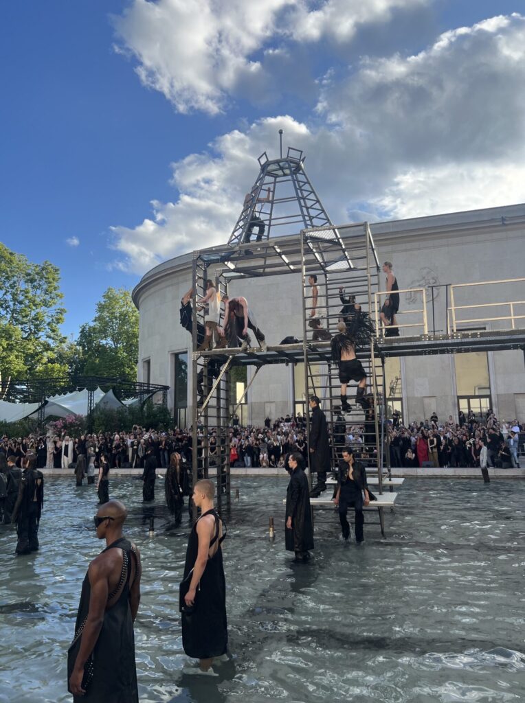 Models descend into the fountain pool at the rear courtyard of the Palais de Tokyo during the Rick Owens SS26 Temple men's show, beneath four stories of dark iron scaffolding.