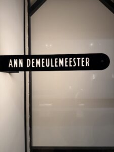 An illuminated black blade sign reading ANN DEMEULEMEESTER, mounted beside the black-framed glass of the new Paris store at the Palais Royal.