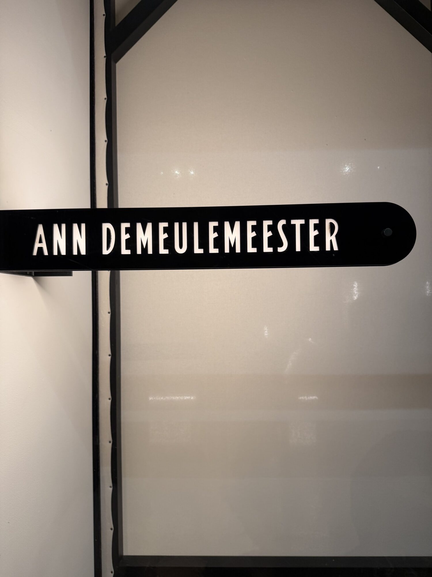An illuminated black blade sign reading ANN DEMEULEMEESTER, mounted beside the black-framed glass of the new Paris store at the Palais Royal.