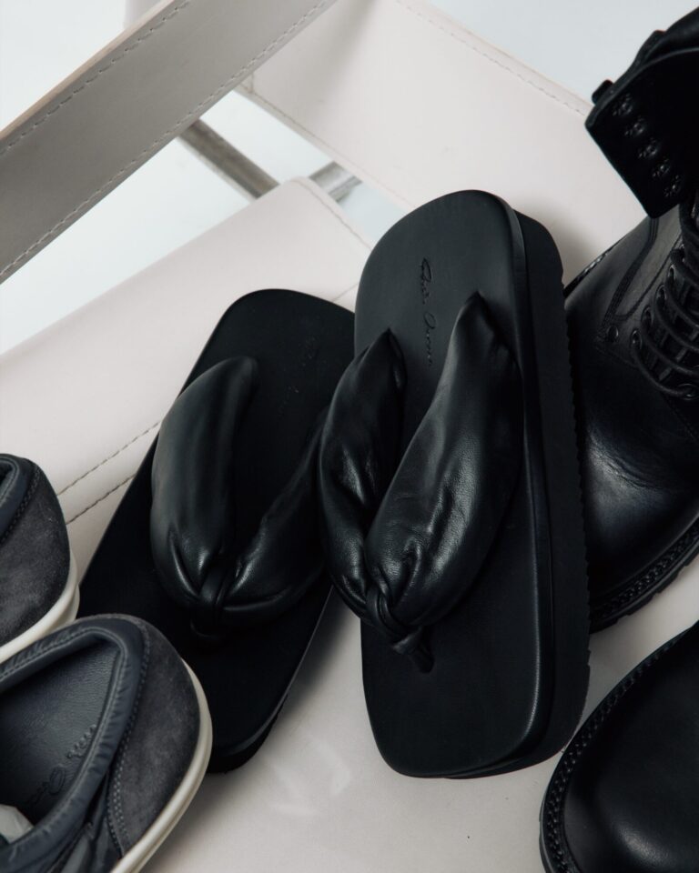 Several pairs of black shoes gathered on a white leather chair: padded leather thong sandals, a lace-up boot, and low suede sneakers.