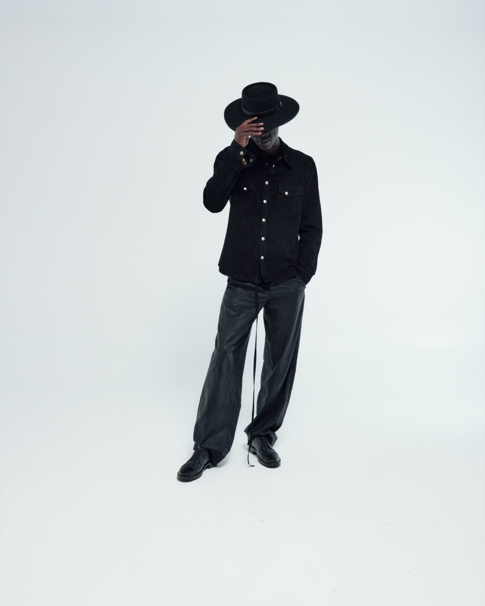 A model in a black wide-brim hat, black snap-front shirt, and waxed wide-leg trousers, photographed against a pale grey studio backdrop.