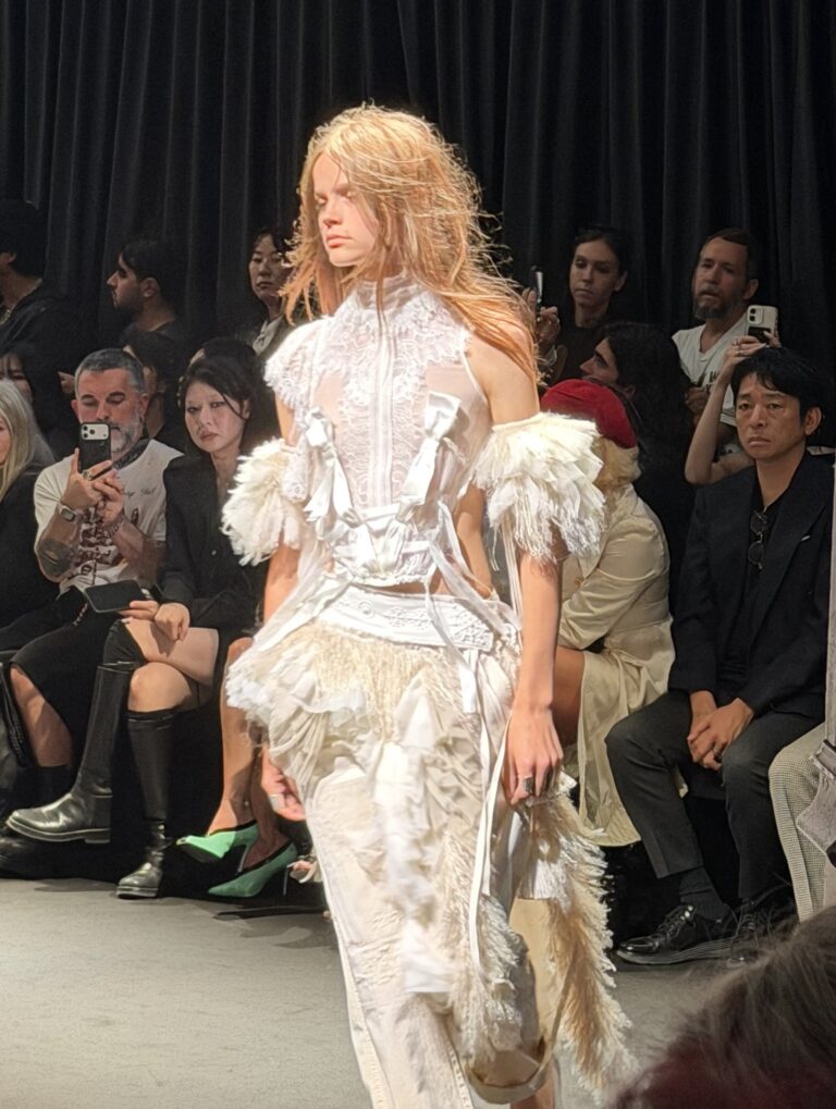 A model with long loose hair in a high-necked cream corset top with feathered sleeves, over a long pale distressed skirt, walking between seated rows.