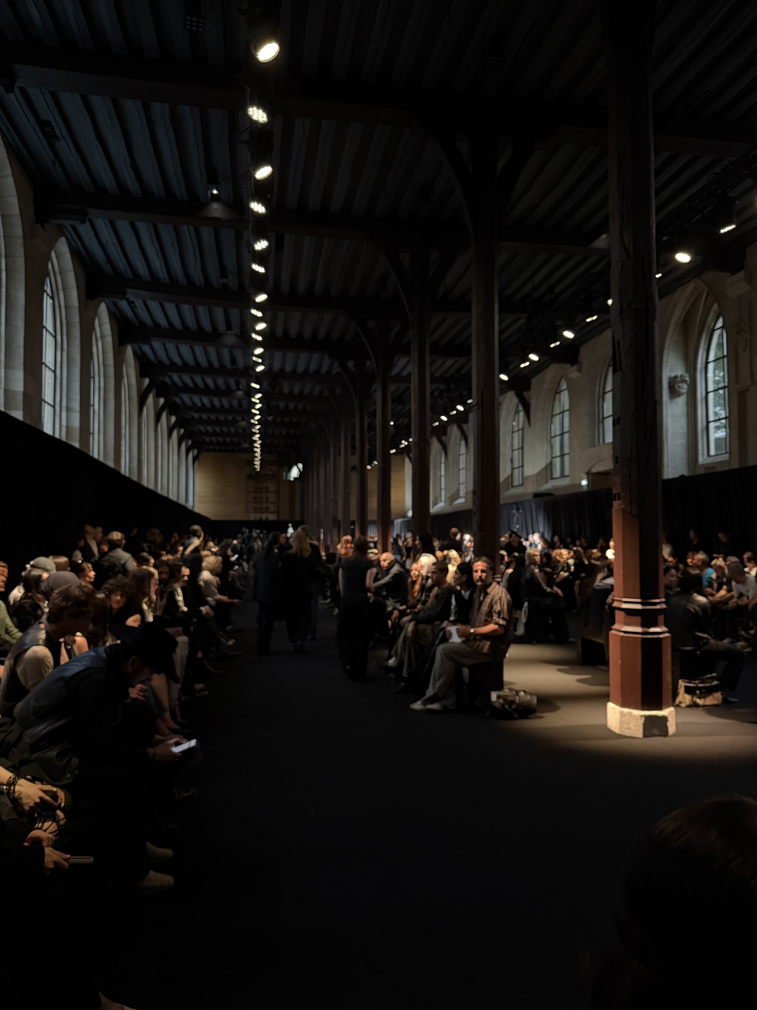 The long vaulted hall before the show, timber roof trusses and stone columns above a dark runway, with the audience seated along both sides.