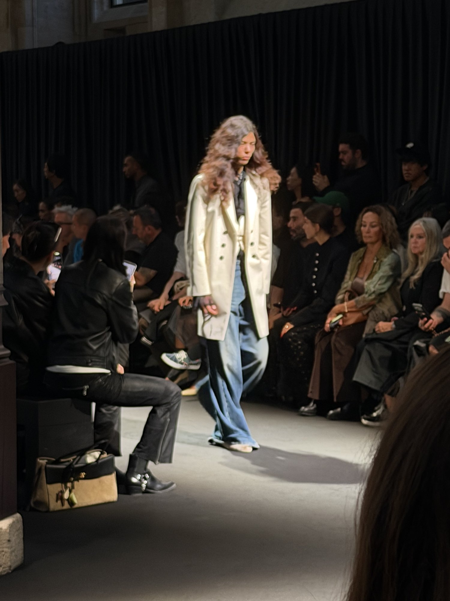 A model in a long cream coat worn open over faded wide-leg denim, walking the runway past the front row.