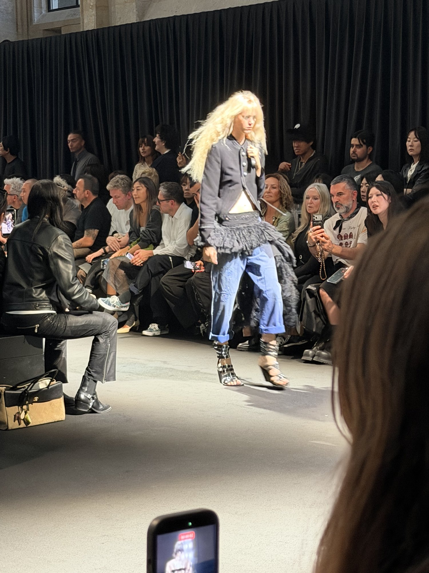 A model with long platinum hair in a grey tailored jacket over blue jeans and a grey fringed skirt, in strappy heels.