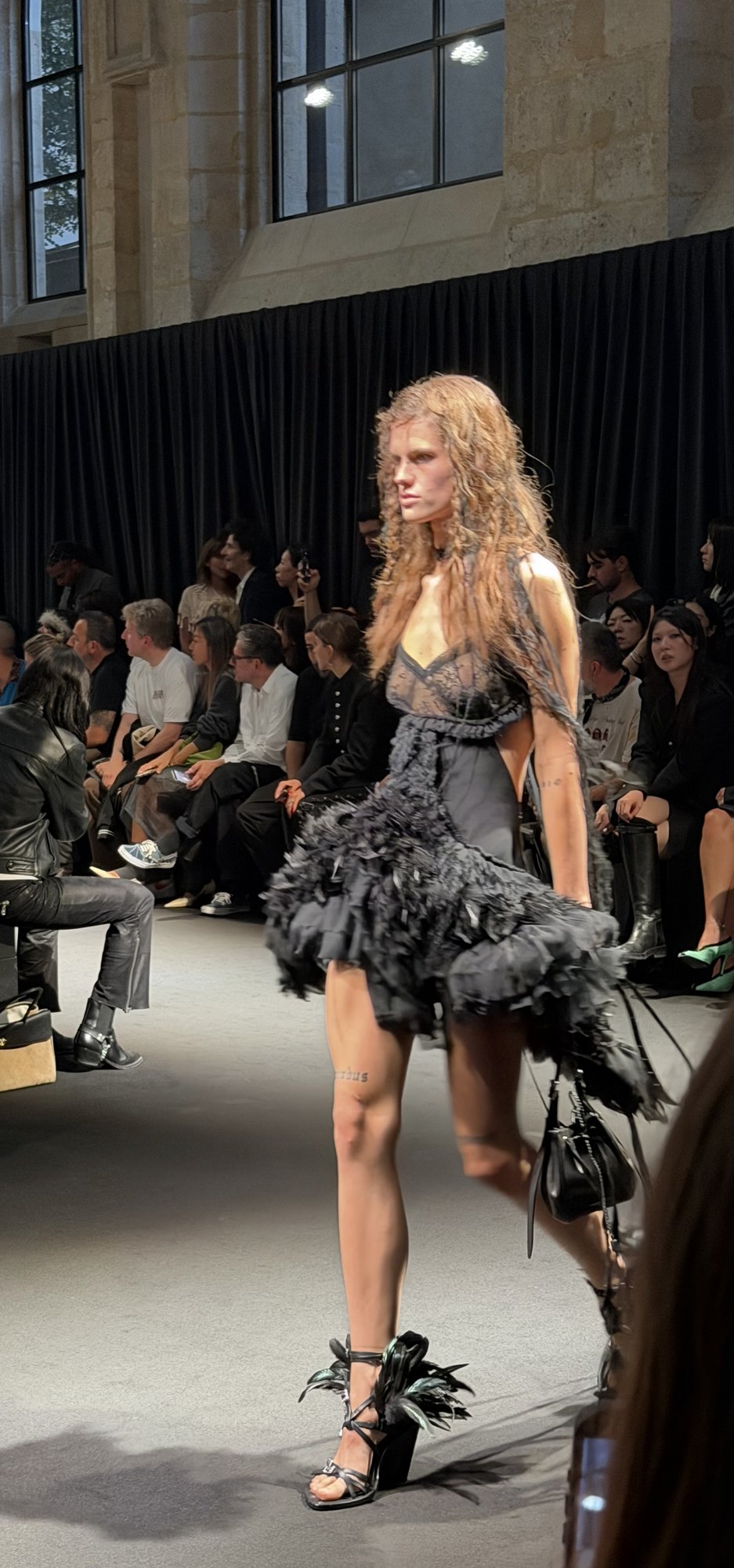 A model in a short dark ruffled and feathered dress with a tightly defined waist, in strappy heels, passing the seated audience.