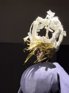 A model in profile under a white padded crown of raw-edged quilted fabric, with stiff gold strands radiating from beneath it like spun hair, over a bulbous paneled blue denim shoulder.