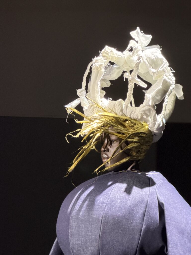 A model in profile under a white padded crown of raw-edged quilted fabric, with stiff gold strands radiating from beneath it like spun hair, over a bulbous paneled blue denim shoulder.