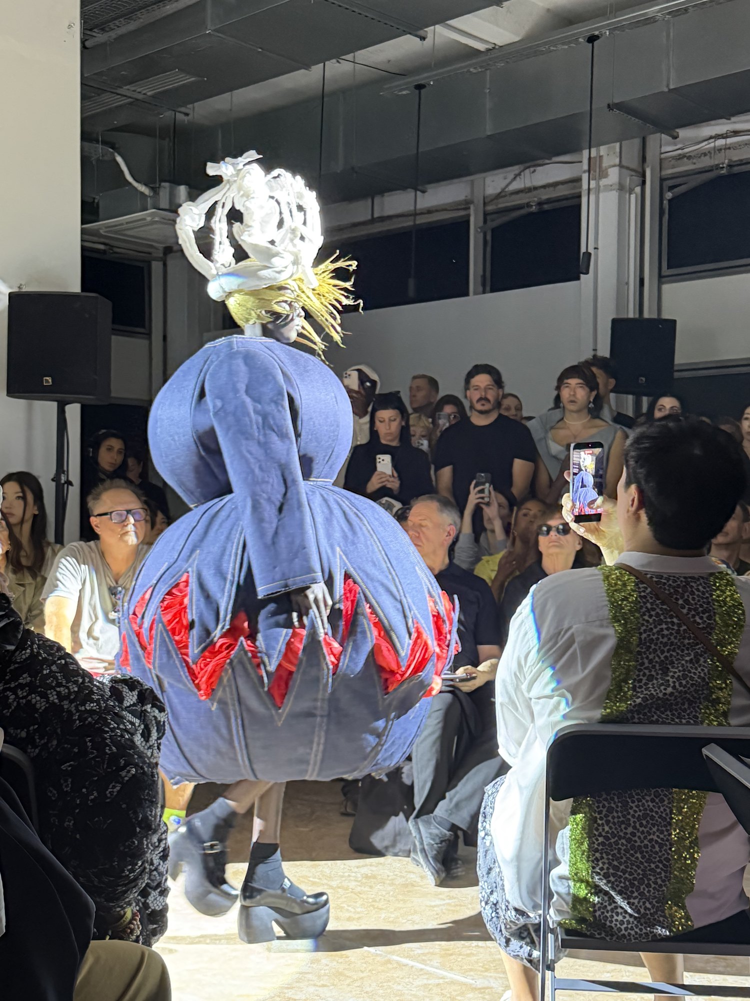 Seen from behind on the runway, a model in a rounded pale blue denim garment with red zigzag shapes at the hem, a white crown above, the seated audience watching from both sides.
