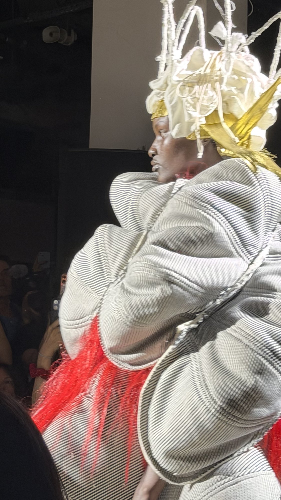A model under a white padded crown threaded with gold, wearing a black and white ticking stripe quilted into bulbous panels, with long red threads spilling from the hem.