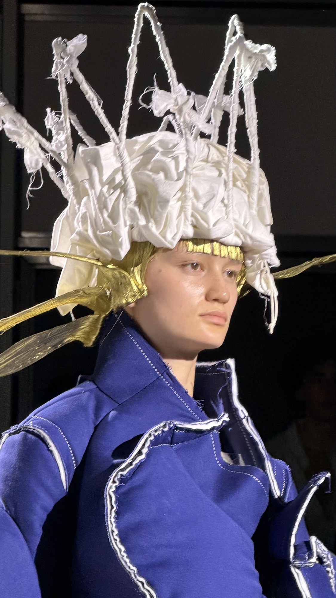 A tall white crown built from padded fabric spikes, gold visible beneath its brim, above an indigo denim garment with contrast topstitching.