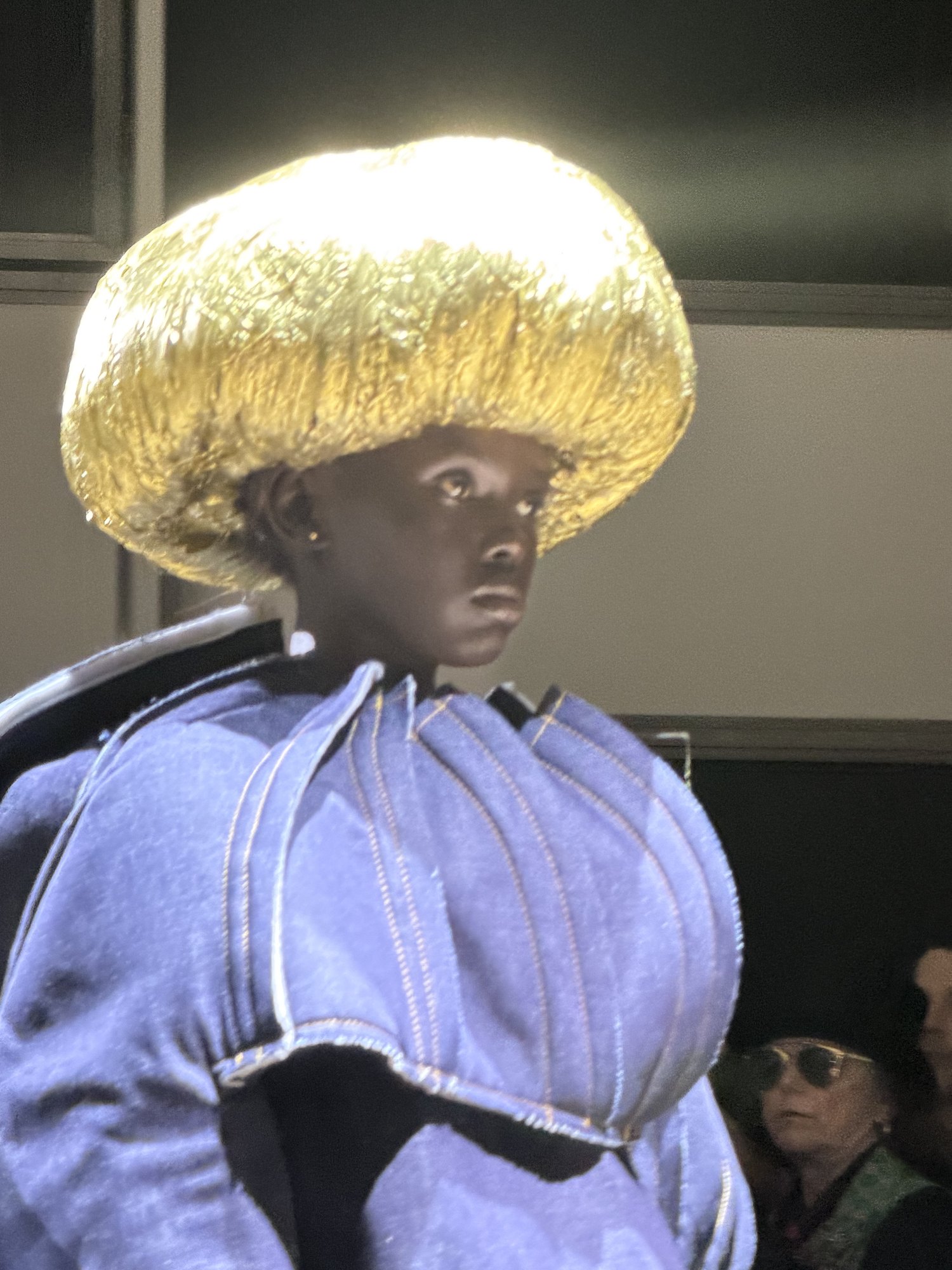 A large domed gold headpiece with a combed, lacquered texture sitting above the brow like a crown, over a bulbous pale blue denim garment with contrast topstitching.