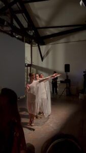 Three dancers in sheer pale layered dresses standing close together with their arms extended to one side, in a high white room with exposed dark timber trusses and a speaker on a stand.