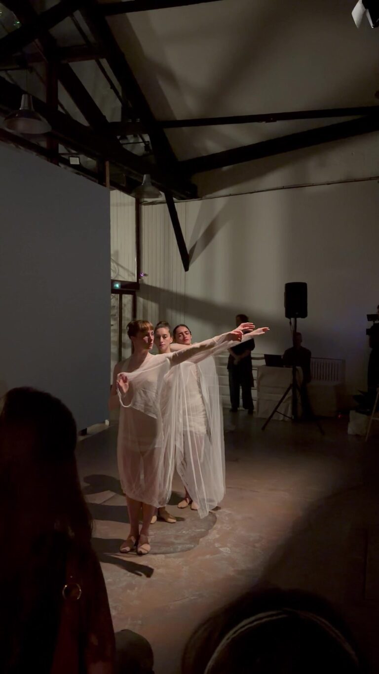 Three dancers in sheer pale layered dresses standing close together with their arms extended to one side, in a high white room with exposed dark timber trusses and a speaker on a stand.