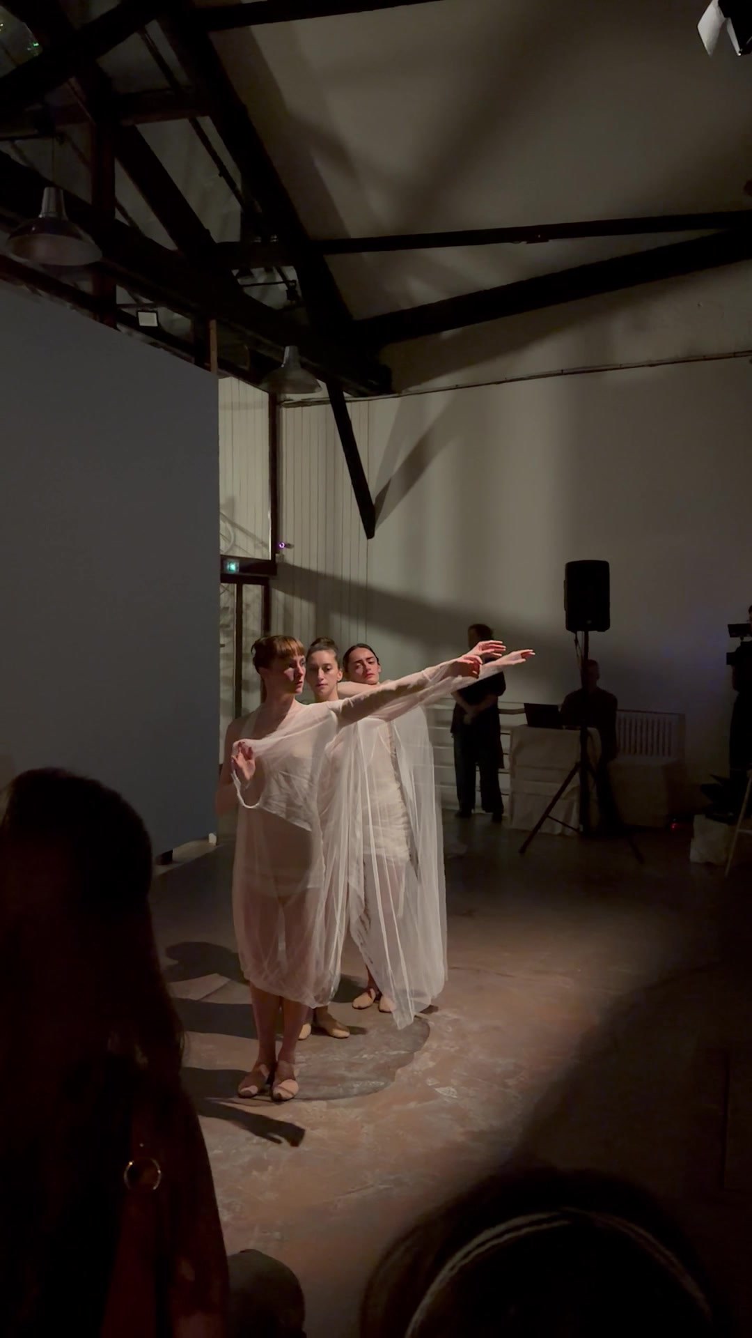 Three dancers in sheer pale layered dresses standing close together with their arms extended to one side, in a high white room with exposed dark timber trusses and a speaker on a stand.