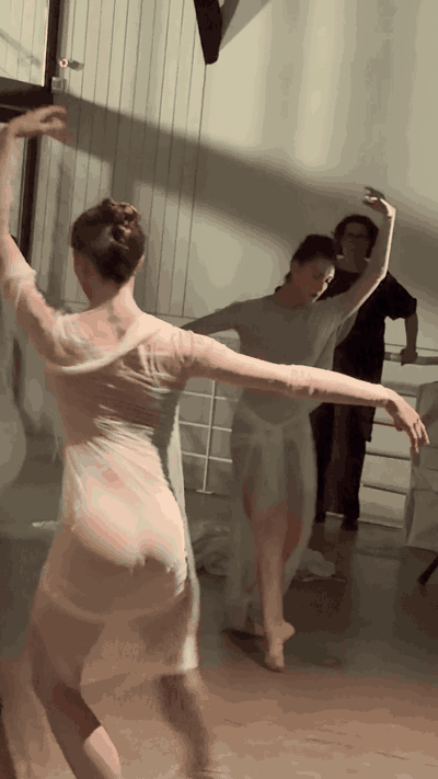 An animated clip of a dancer in a sheer draped pale dress turning with one arm raised, the long skirt lifting as she moves, with other dancers behind her.