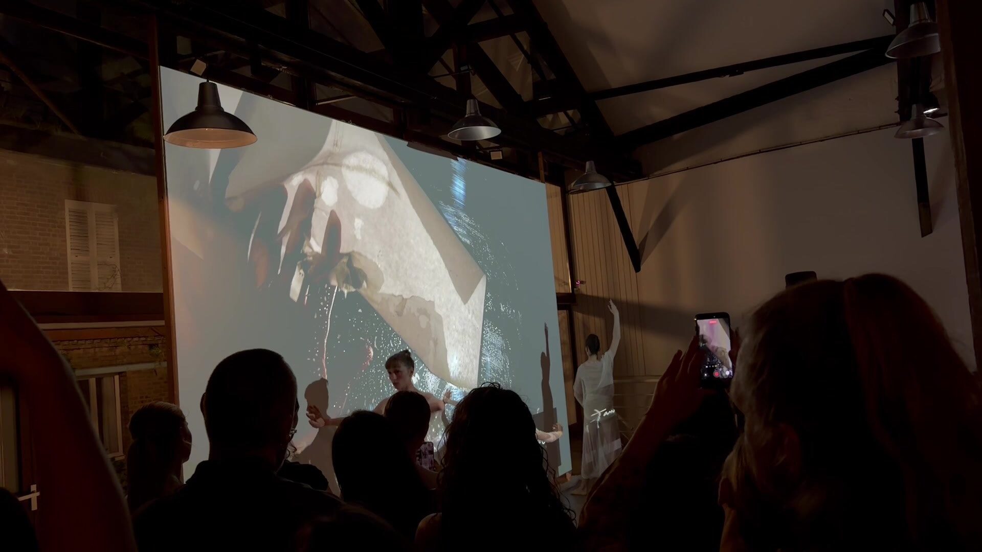 A large tilted projection screen showing a pale figure and draped white fabric beside water, above the silhouetted heads of guests, framed by black scaffolding.