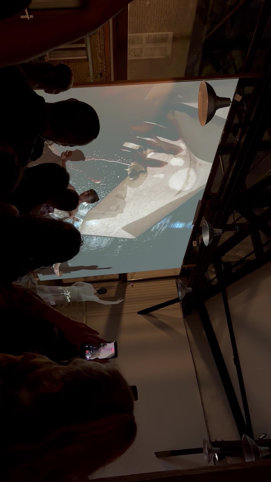 A large tilted projection screen showing a pale figure and draped white fabric beside water, above the silhouetted heads of guests, framed by black scaffolding.