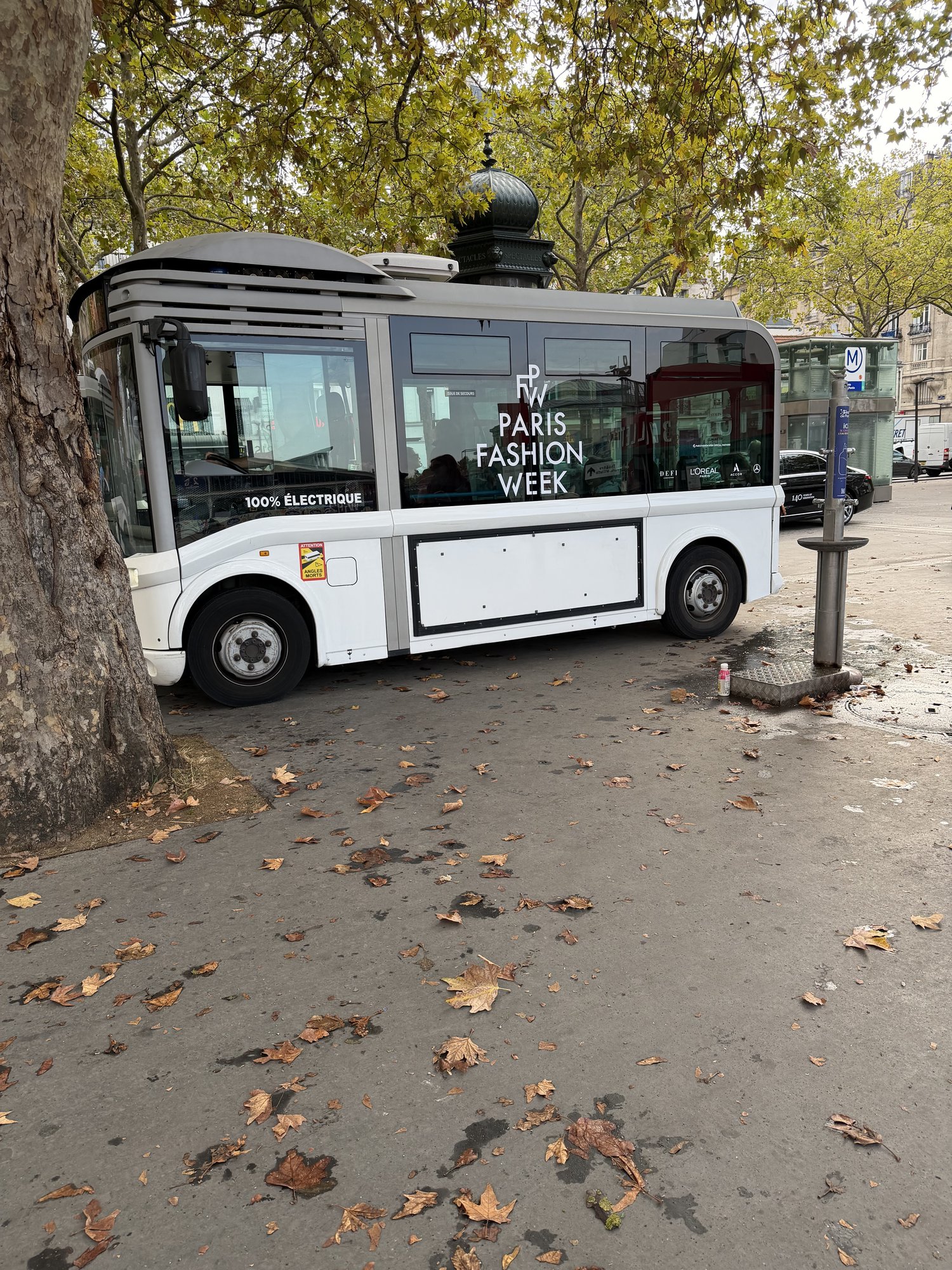 A white electric shuttle marked Paris Fashion Week parked under plane trees, fallen autumn leaves on the pavement.