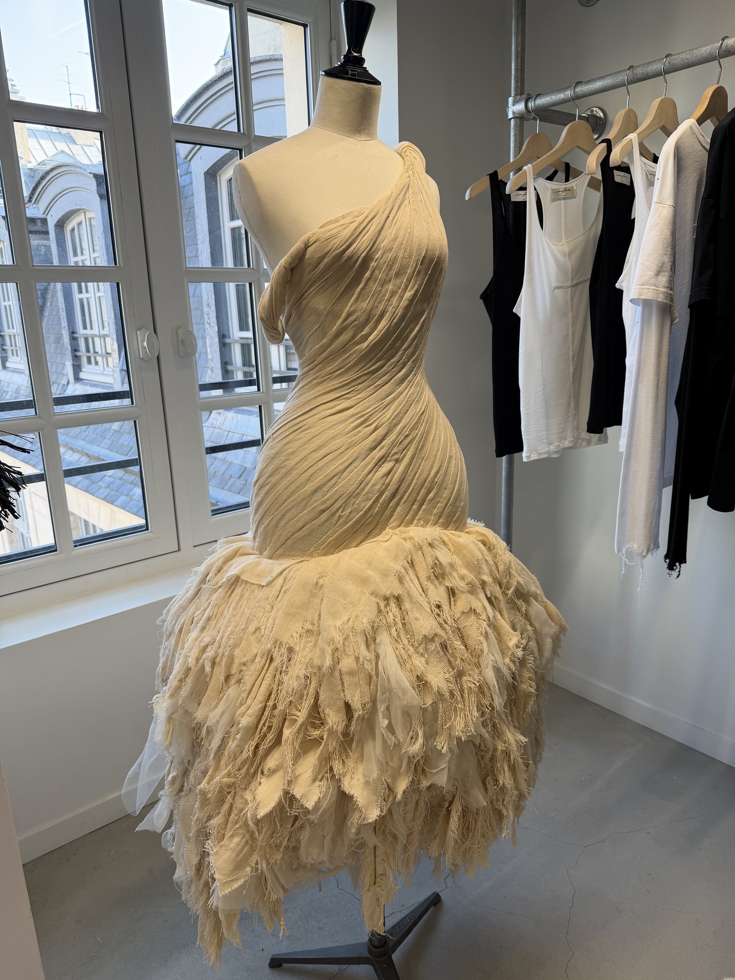 A one-shoulder dress on a dress form by a showroom window, cream fabric twisted tightly over the body and breaking into a skirt of frayed, feather-like layers.