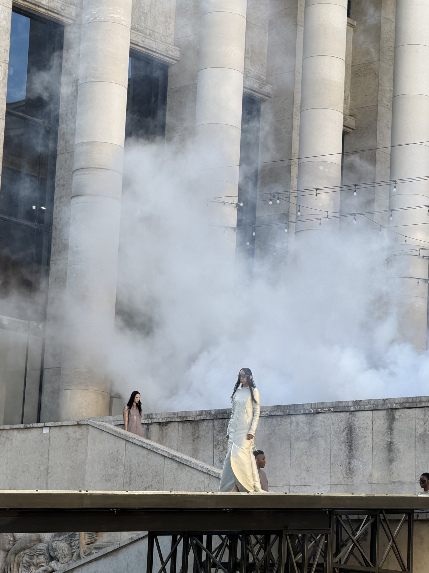 Mist billows between the stone columns of the Palais de Tokyo as a model in a long, pale, high-necked dress walks the Rick Owens runway, another descending the steps behind her.