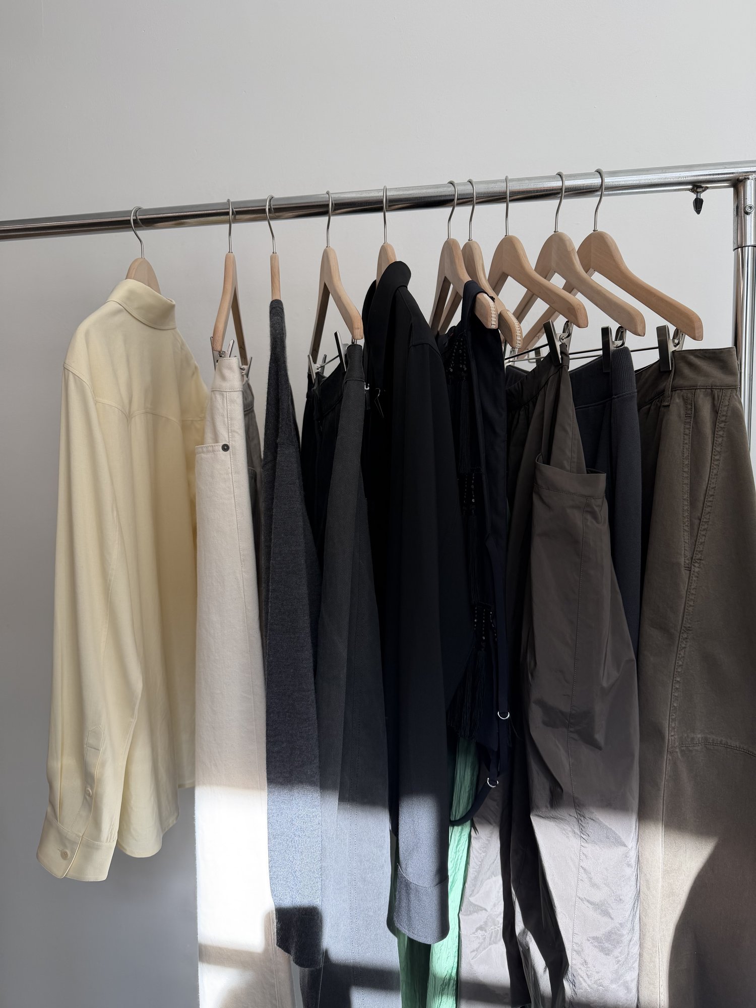 A rail of clothes on wooden hangers in a sunlit showroom: a pale yellow shirt, cream trousers, grey knitwear, black pieces, and olive trousers.