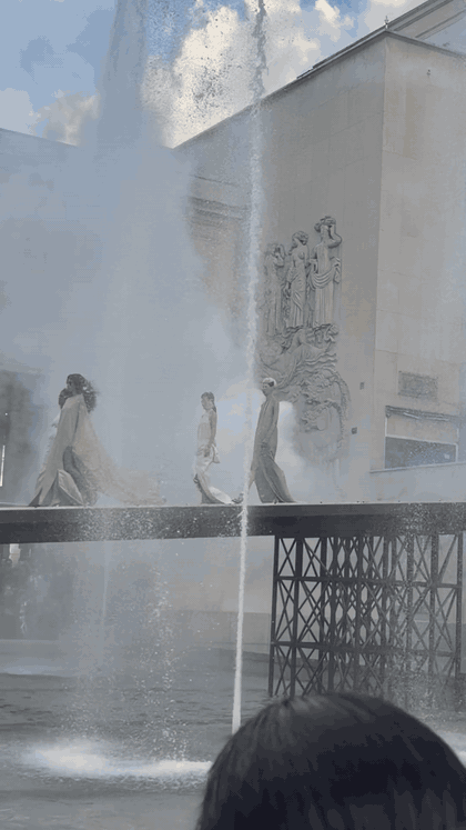 Models in pale trailing garments cross the reflecting pool as fountains spray at the Palais de Tokyo